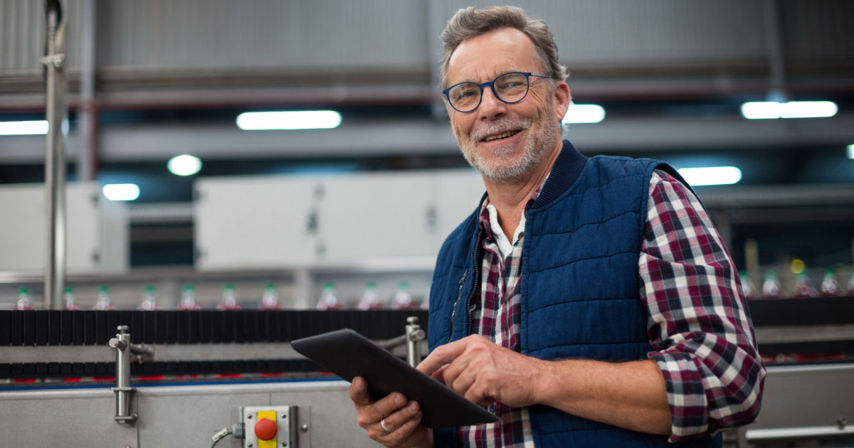 Older factory worker inspecting electrical machinery with tablet in hand