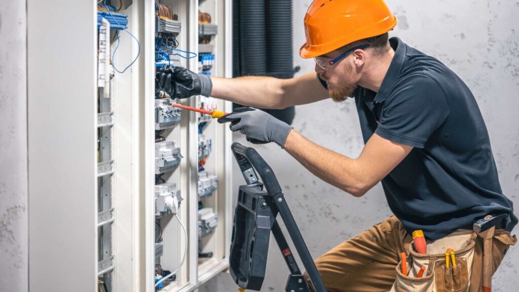 Electrician with orange hard hat and utility pants looks at wires in a wall box 